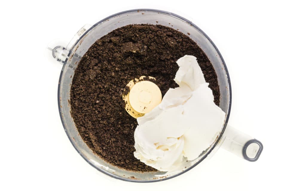Vegan cream cheese has been added to a food processor bowl with chocolate cookie crumbs.