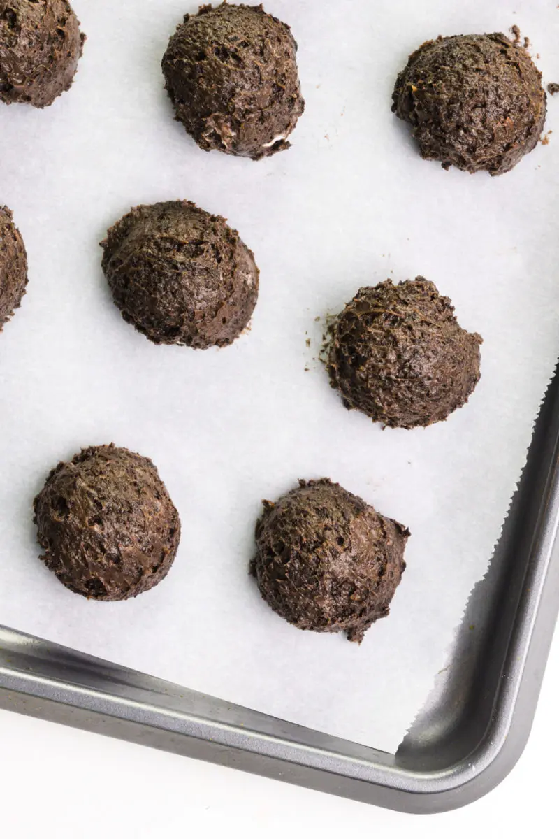 Balls of chocolate dough are lined up on a baking sheet lined with parchment paper. 