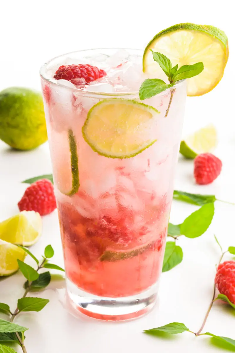 A glass holds a red beverage with slices of lime, mint, and raspberries both in the glass and around the glass too.