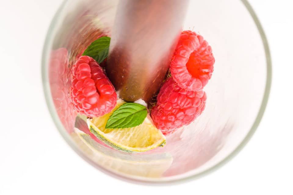 A cocktail muddler is in a glass with fresh raspberries, mint, and lime wedges.