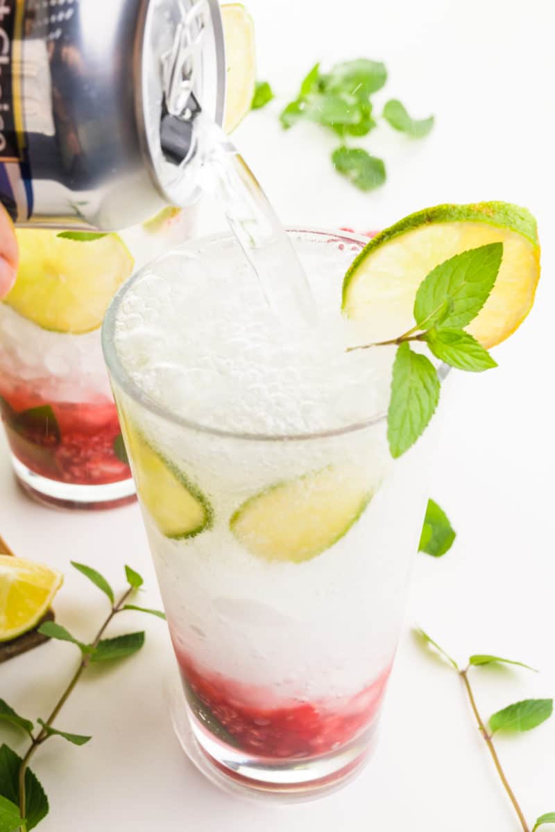 A hand holds a can of club soda and is pouring it into a glass with crushed ice and limes, and a raspberry mixture in the bottom of the glass.