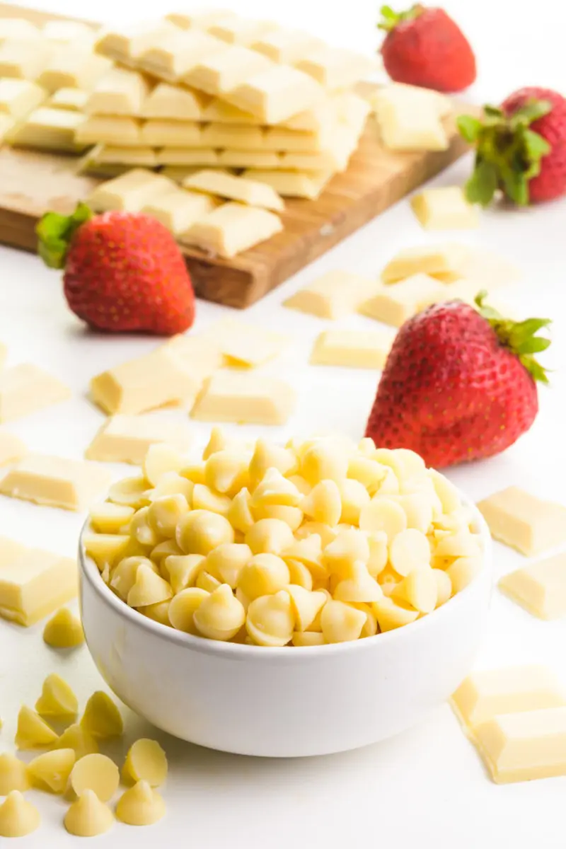 A bowl of white chocolate chips sits in front of and around fresh strawberries and more of the chips on the table. There's a cutting board in the background with white chocolate bars.