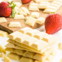 A stack of several vegan white chocolate bars sit in front of a wooden cutting board with more of the chocolate chunks and fresh strawberries.