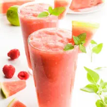 Two glasses full of watermelon slushies have raspberries and mint leaves around them. There are watermelon wedges as garnishes in the drink and also sitting behind the drinks too.