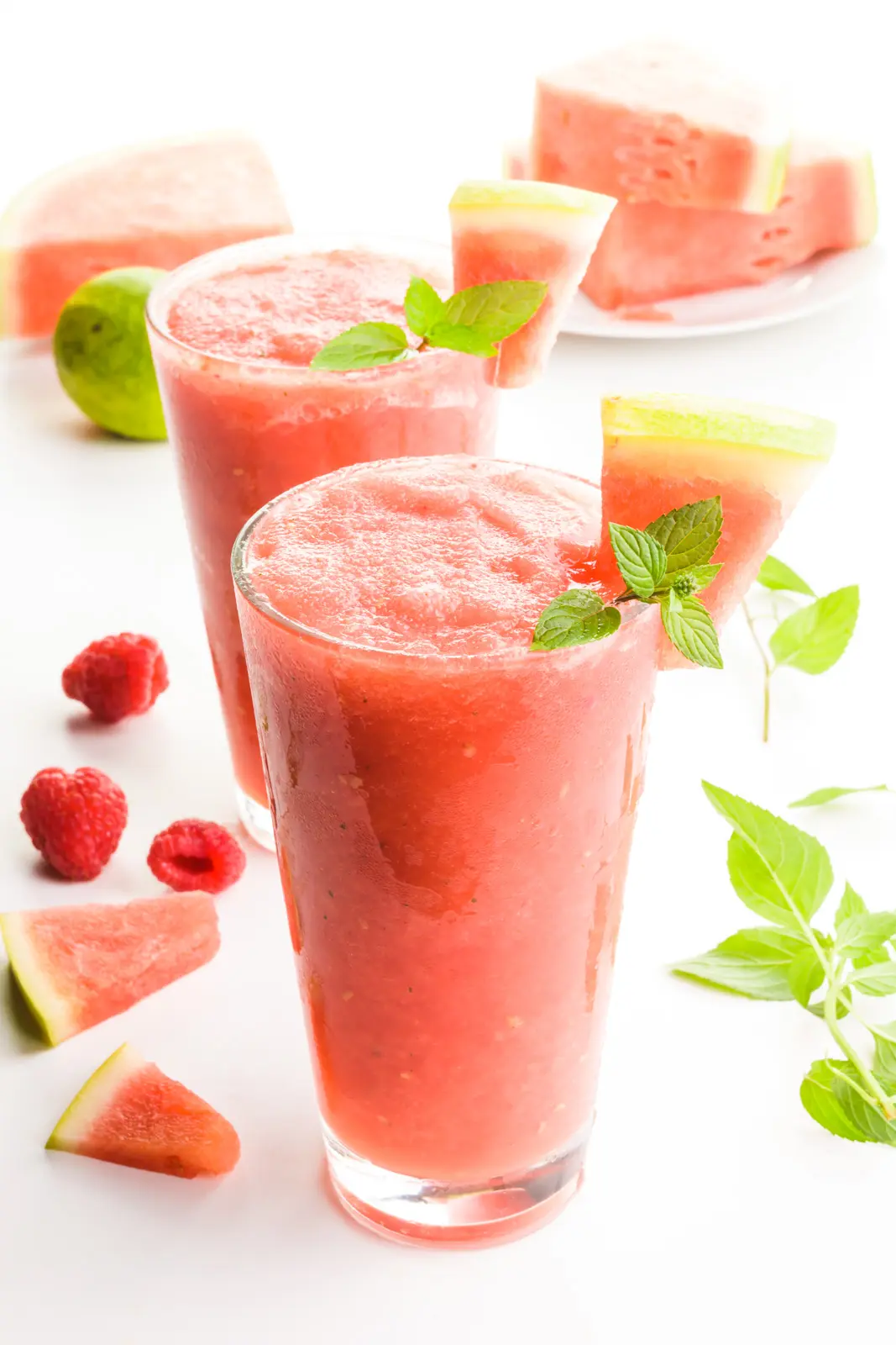 Two glasses full of watermelon slushies have raspberries and mint leaves around them. There are watermelon wedges as garnishes in the drink and also sitting behind the drinks too.