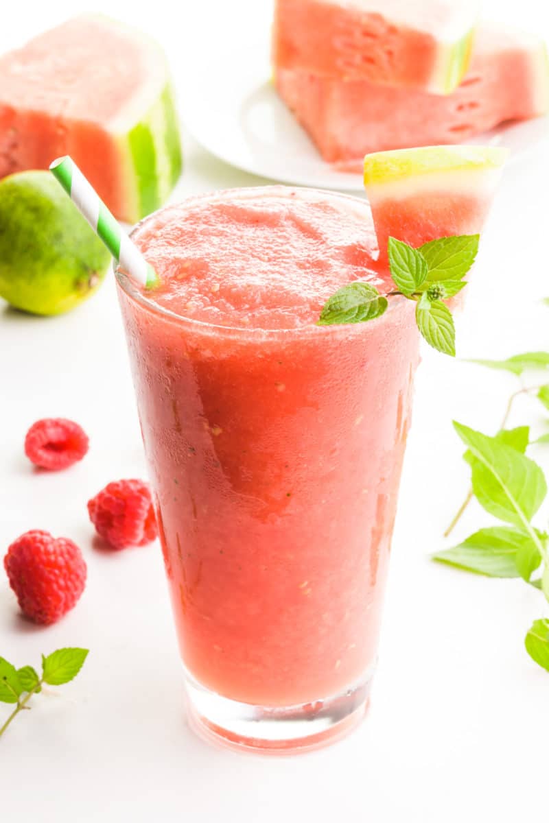 A glass holds a pink beverage with watermelon slices as garnish. There's fresh raspberries, mint leaves, and watermelon slices around it.