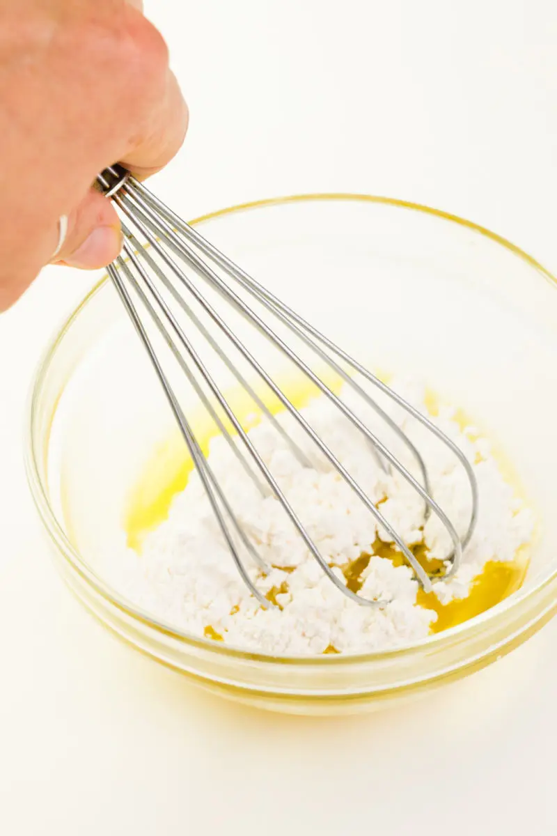 A hand holds a whisk stirring a bowl of melted cocoa butter and powdered sugar.