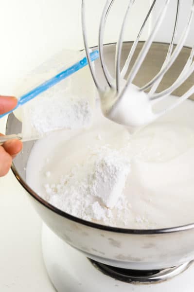 Powdered Sugar is being added to whipped cream in a stand mixer bowl.
