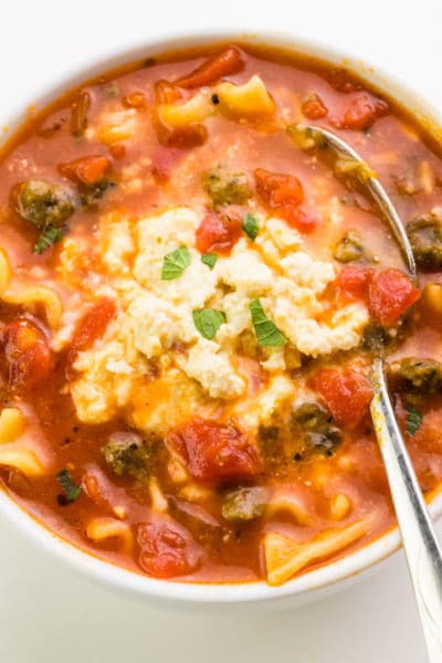 A spoon is in a bowl of tomato-based soup, with lasagna noodles and a creamy sauce on top.