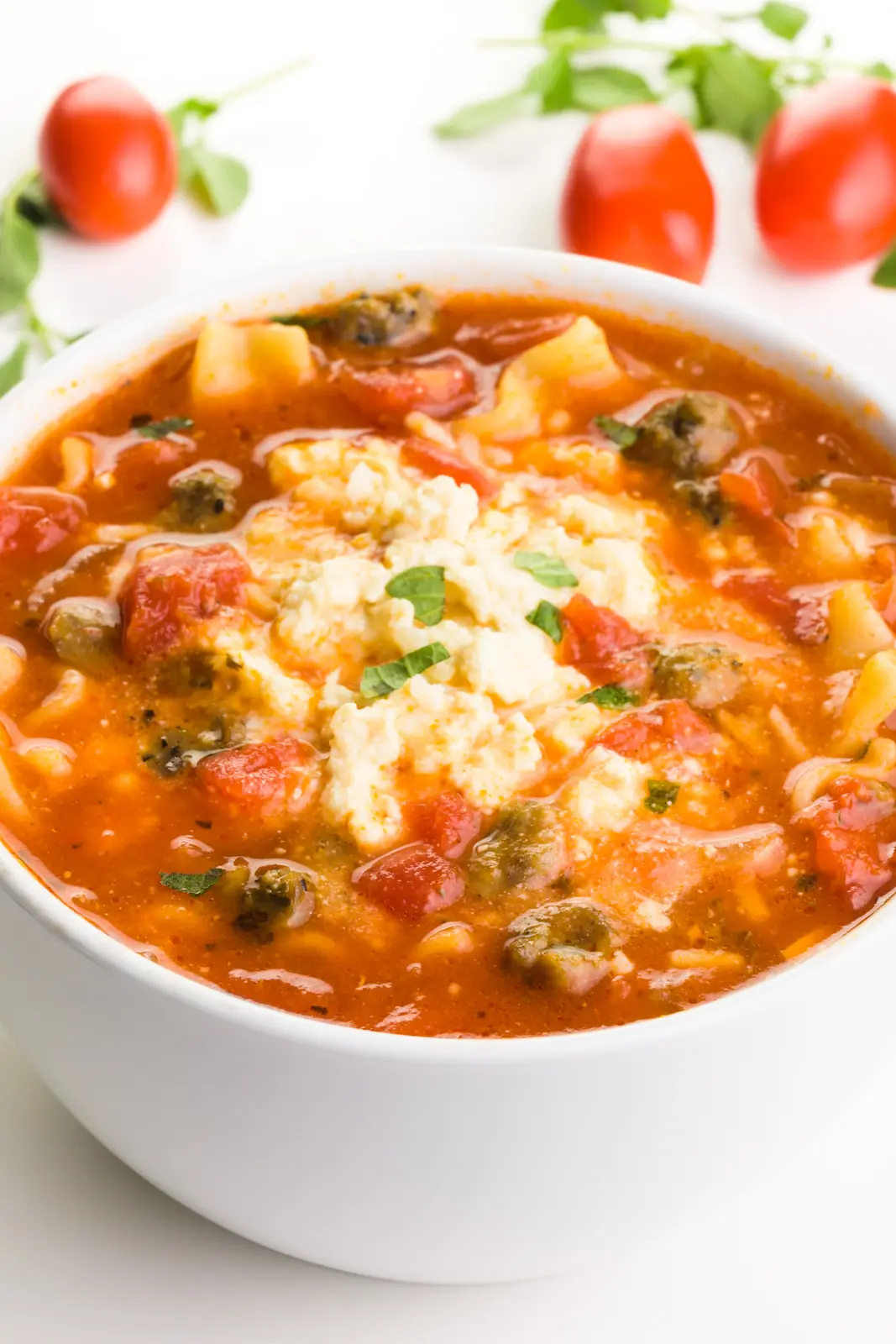 A bowl holds vegan lasagna soup with a creamy sauce on top. There are cherry tomatoes and fresh herbs around it.