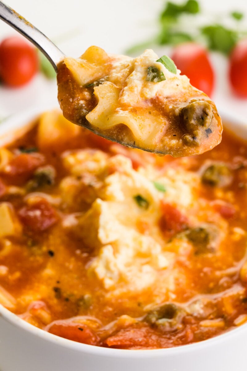 A spoon full of soup hovers over the rest of the bowl. There are cherry tomatoes in the background and fresh herbs.