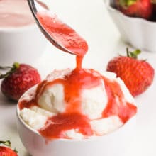 A hand holds a spoon drizzleling strawberry sauce over ice cream. There are fresh strawberries and a bowl of more sauce in the background.