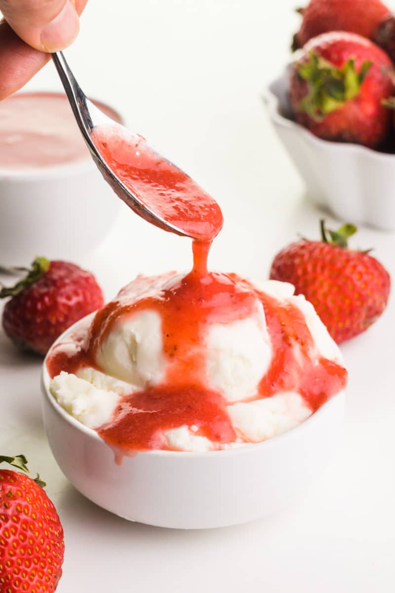 A hand holds a spoon drizzleling strawberry sauce over ice cream. There are fresh strawberries and a bowl of more sauce in the background.