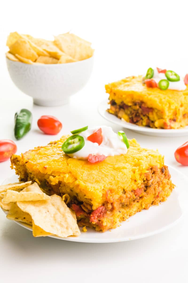 Two plates hold slices of taco cornbread casserole. There's a bowl of tortilla chips behind it.