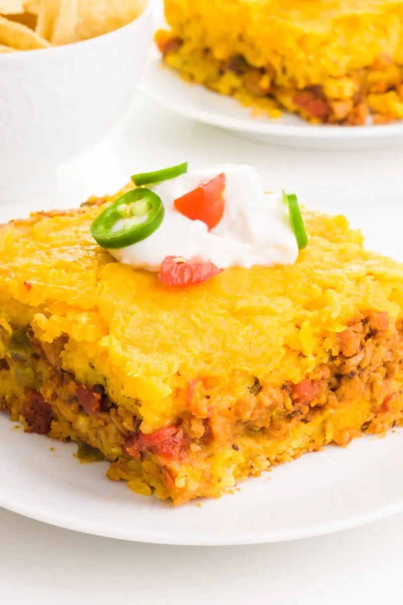 A close-up of a Mexican cornbread casserole slice on a plate. It's topped with sour cream, chopped tomatoes, and chopped jalapeños.. 