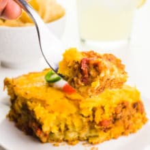A hand holds bite of casserole, hovering over the rest of the slice. There's a bowl to tortilla chips and a margarita in the background.
