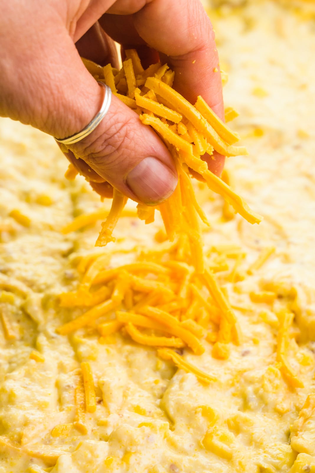 A hand holds cheese and is spreading it across the top of an unbaked casserole.