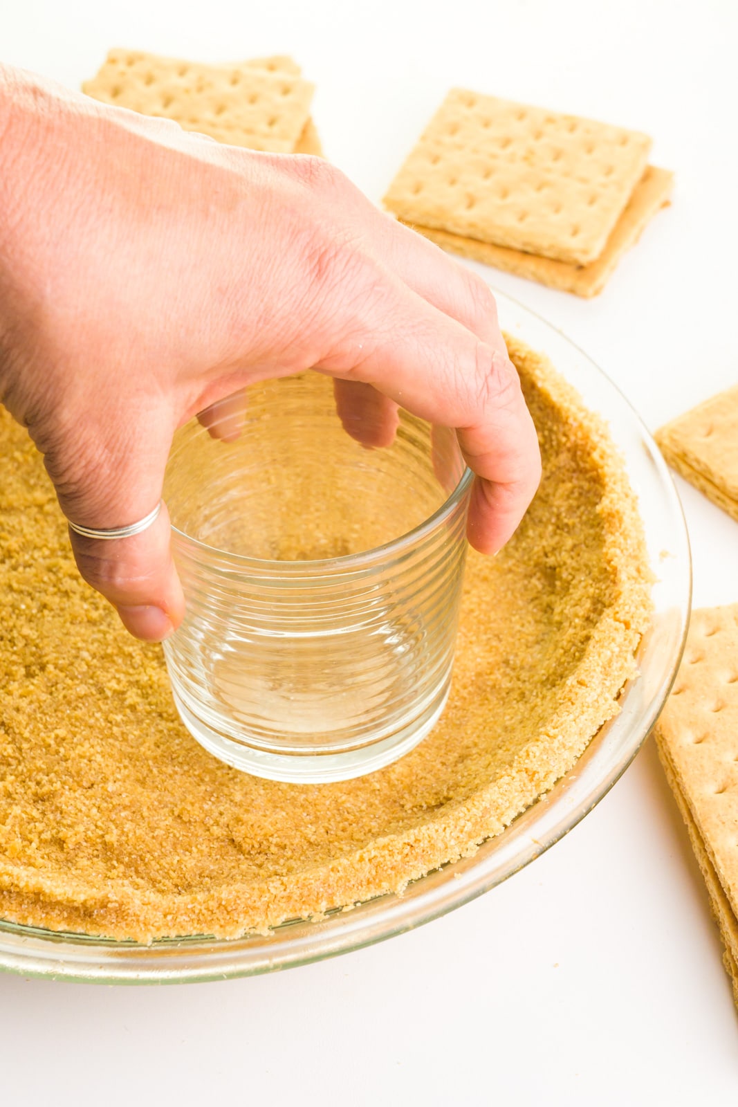 A hand holds a glass, using it to press crumbs into a pie pan.