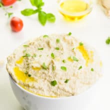 A bowl holds vegan ricotta with fresh herbs on top. There are cherry tomatoes and herbs behind it, along with a bowl of olive oil.