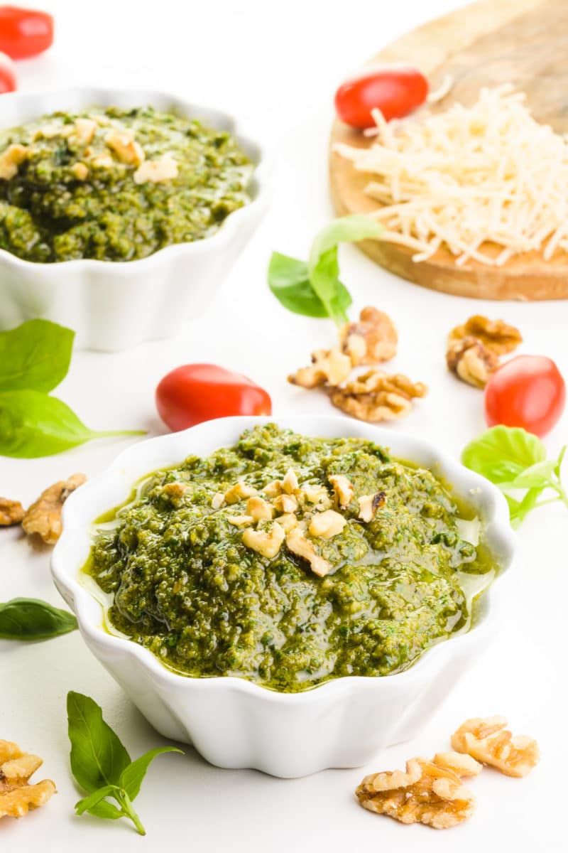 Two bowls hold walnut pesto, topped with chopped walnuts. Around the bowls are cherry tomatoes, basil leaves, and vegan parmesan cheese.