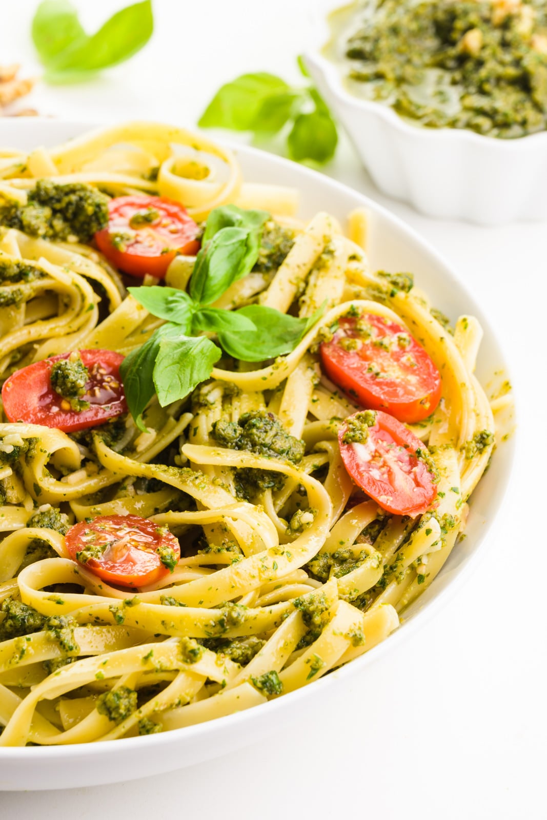 A bowl of pasta has chopped tomatoes and fresh basil leaves on top. Behind it is a bowl of green sauce (pesto) that was added to the pasta as well.
