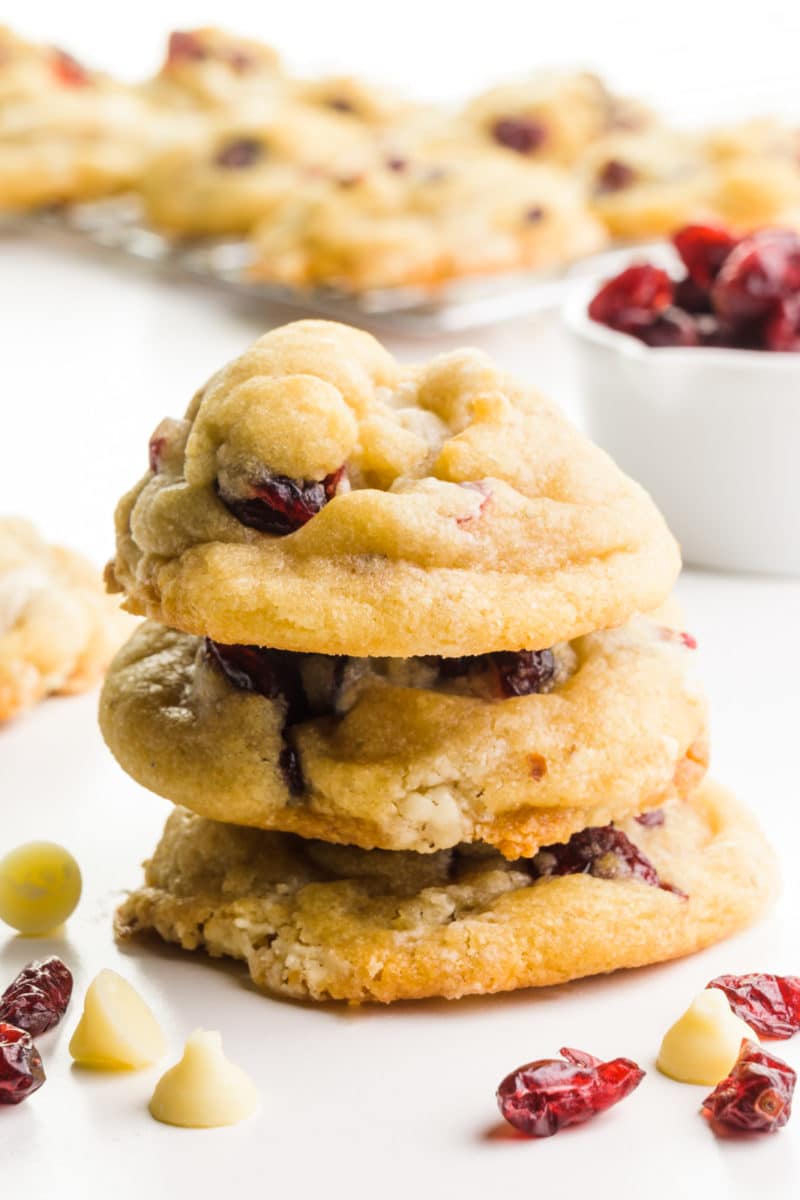 A stack of white chocolate cranberry cookies sits in front of more cookies. There are dried cranberries and white chocoalte chips around them as well.