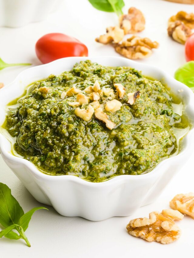 A bowl of walnut pesto has chopped walnuts on top. It's surrounded by walnuts, fresh basil leaves, and cherry tomatoes.