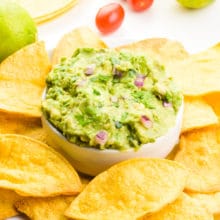 Air fryer tortilla chips are on a plate surrounding a bowl of guacamole. Behind it are fresh lemons, cherry tomatoes, and more tortillas.