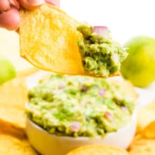 A hand holds a tortilla chip with guacamole on it. There is a bowl with guacamole and more chips behind it.