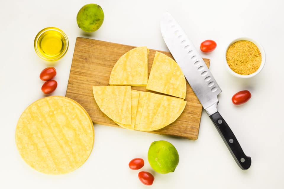 Corn tortillas sit on a cutting board. Some of them have been cut into quarters and they're sitting next to a knife. There are limes, cherry tomatoes, and bowls of spices and olive oil sitting nearby.