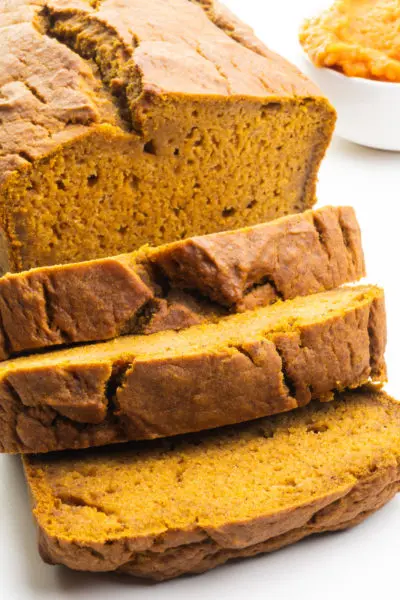 A loaf of vegan pumpkin bread has several slices cut. There's a bowl of Pumpkin Puree next to the loaf.