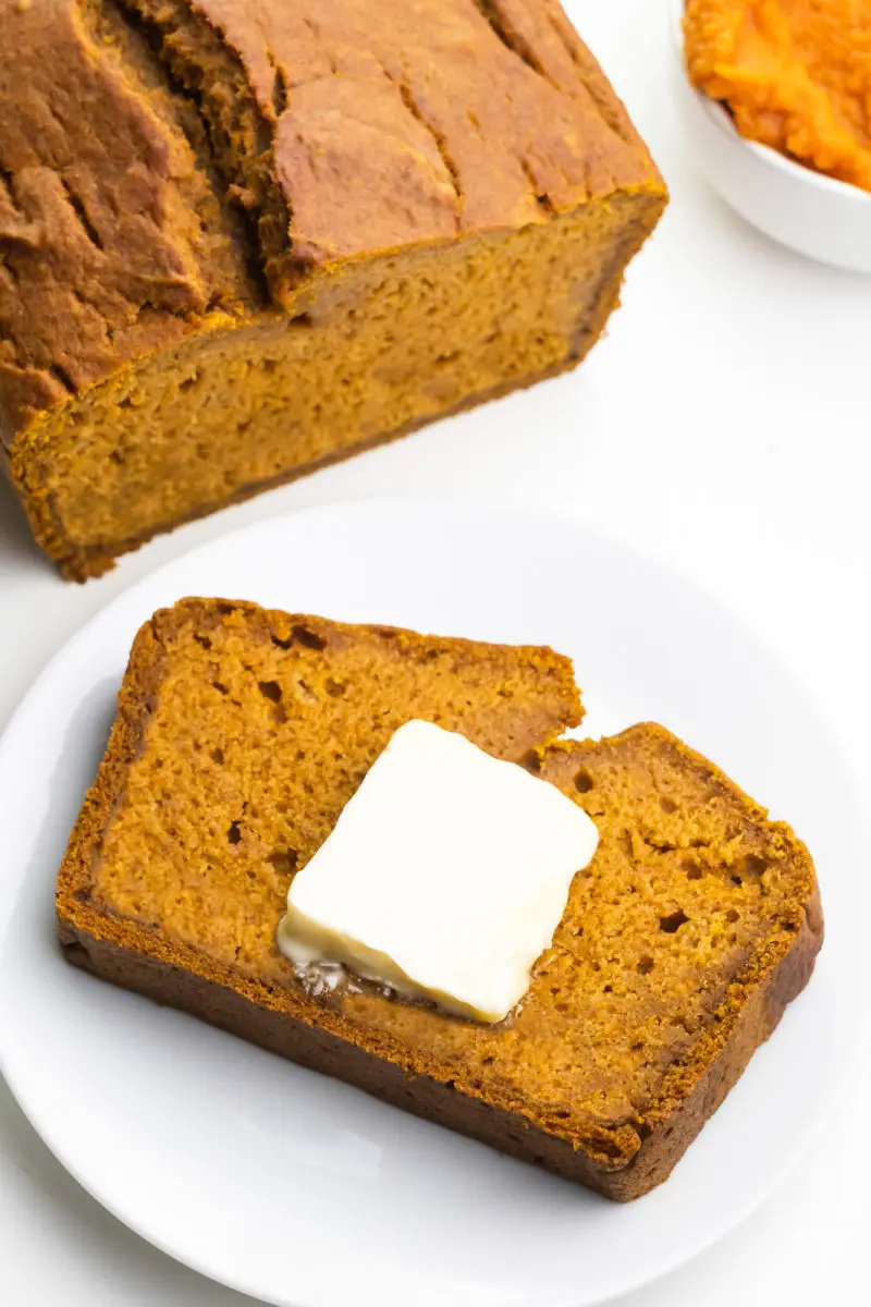 Looking down on a slice of pumpkin bread on a plate. There's a pat of butter on the slice. The rest of the loaf sits nearby alongside a bowl of Pumpkin Puree.