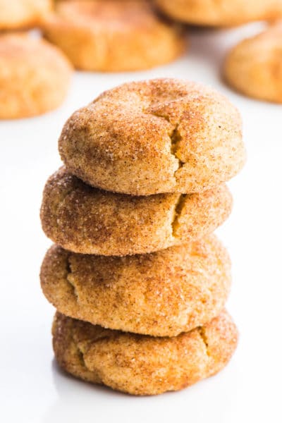 A stack of four snickerdoodles cookies sit one on top of the other, with lots of cinnamon sugar coating. There are several cookies in the background.