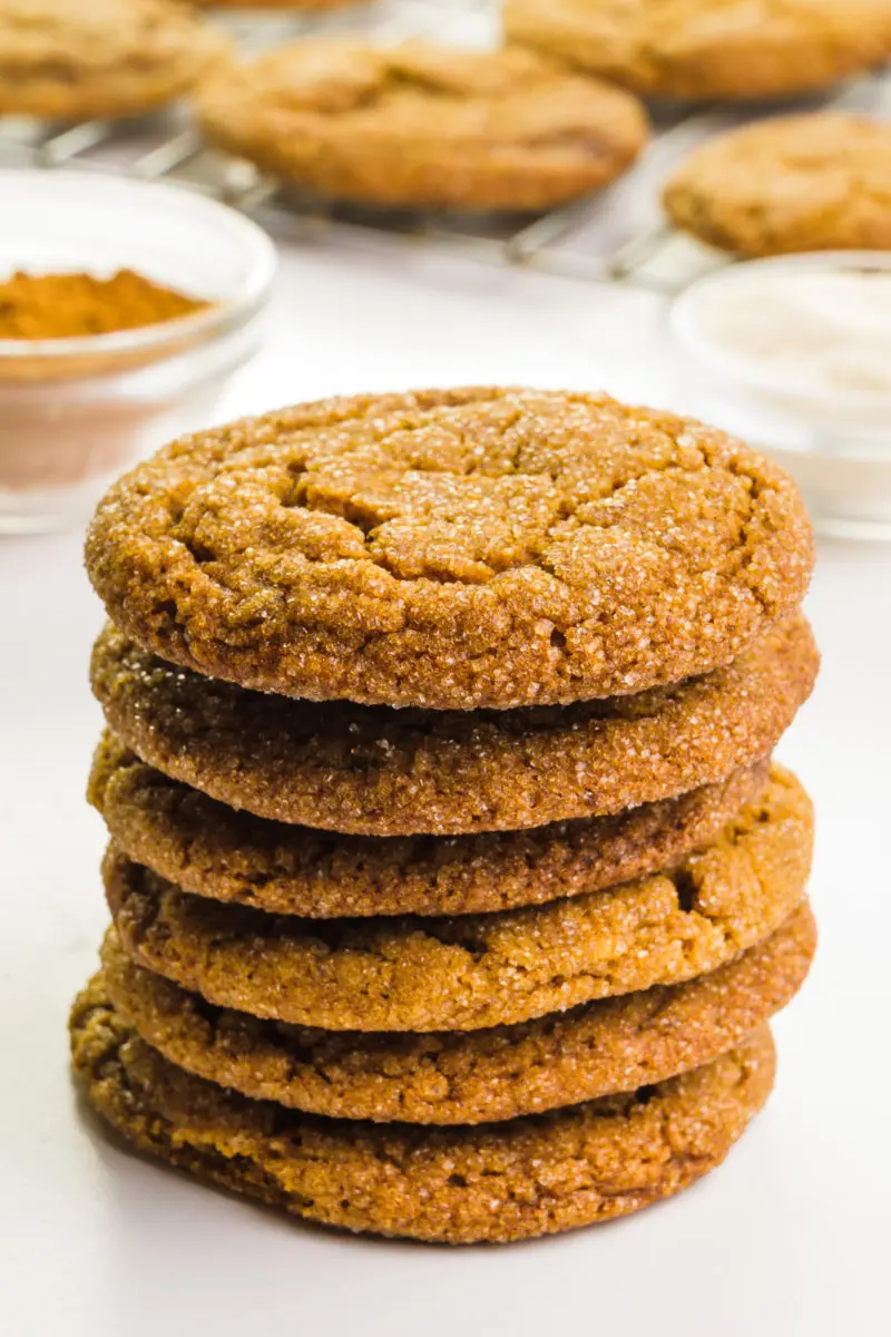 A stack of big soft molasses cookies sits in front of bowls of cinnamon and sugar. There are more cookies on a wire rack in the background.