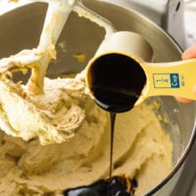 A measuring cup holds molasses and is being poured into a mixing bowl with creamy cookie batter below it.