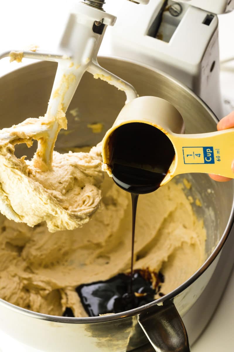 A measuring cup holds molasses and is being poured into a mixing bowl with creamy cookie batter below it.