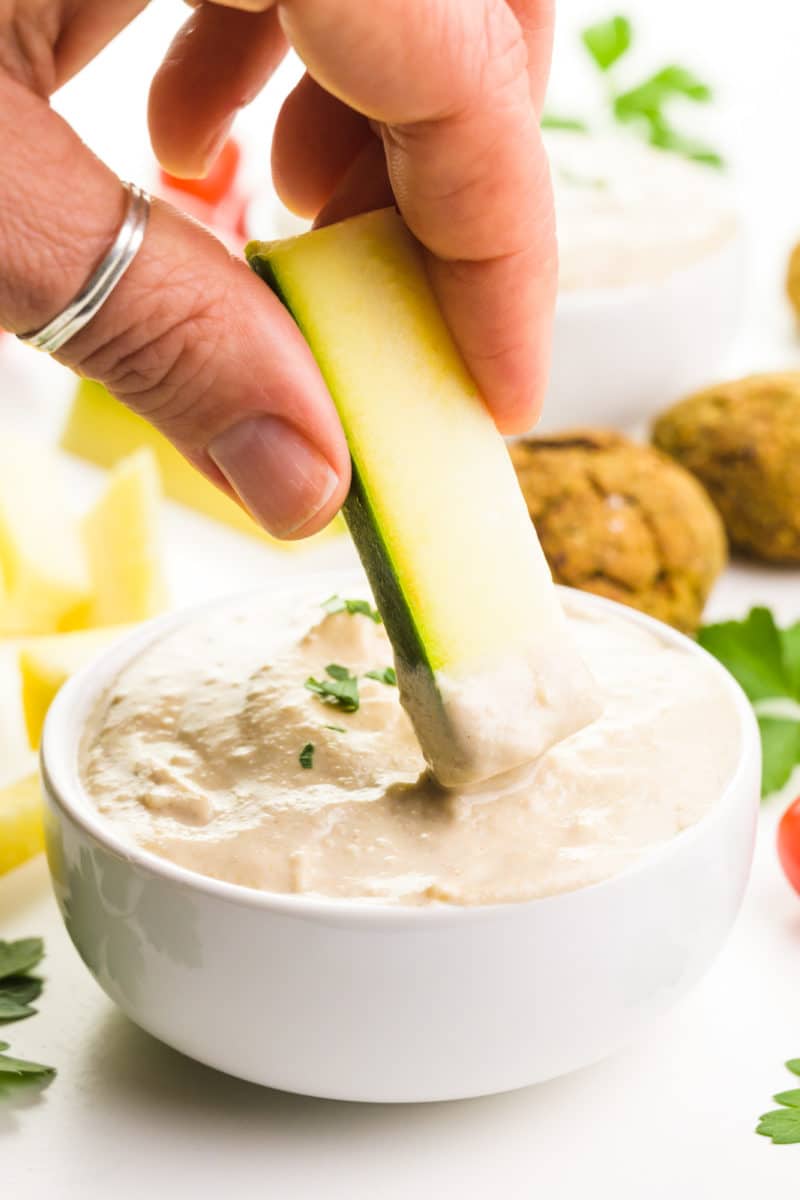 A hand holds a piece of zucchini and is dipping it in a creamy dip. Behind it are fresh herbs and other ingredients for dipping.