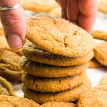 A hand grabs the top cookie in a stack of vegan ginger cookies. There are more cookies in front and behind the stack.