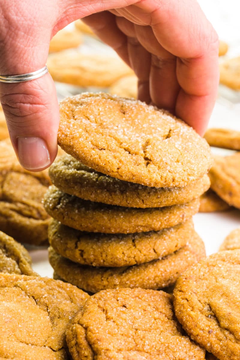 A hand grabs the top cookie in a stack of vegan ginger cookies. There are more cookies in front and behind the stack.