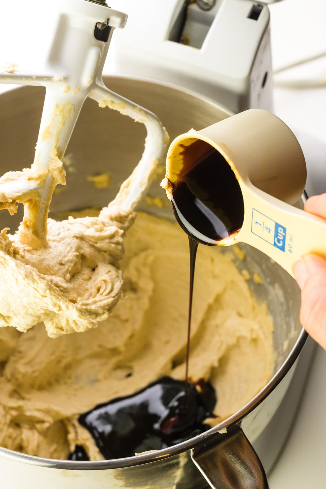 A hand holds a measuring cup pouring molasses into a mixing bowl with cookie batter.