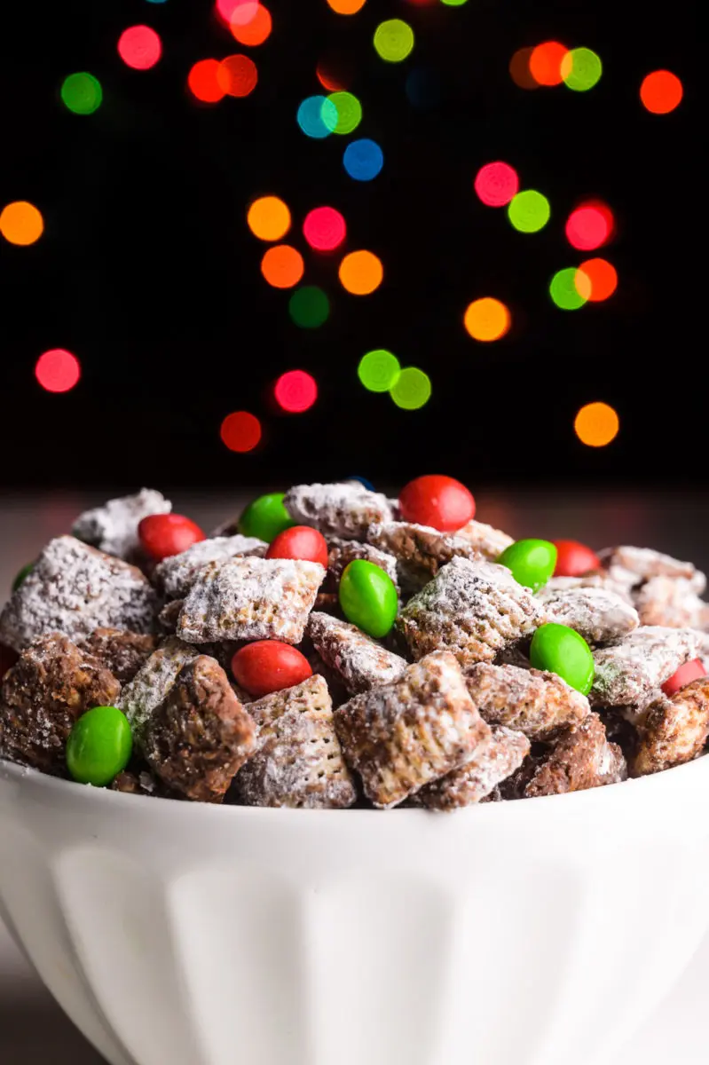 A bowl of chocolate Christmas muddy buddies has a dark background and holiday lights behind it.