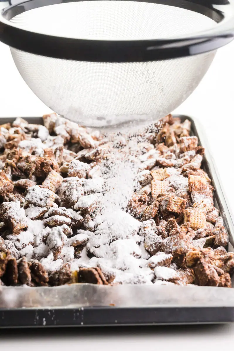 Powdered sugar is being sifted over a pan with chocolate-coated cereal.