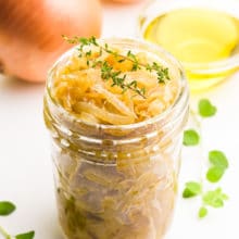 A mason jar holds onion confit with sprigs of thyme on top. There's more herbs around it, and a bowl of olive oil and onions behind it.