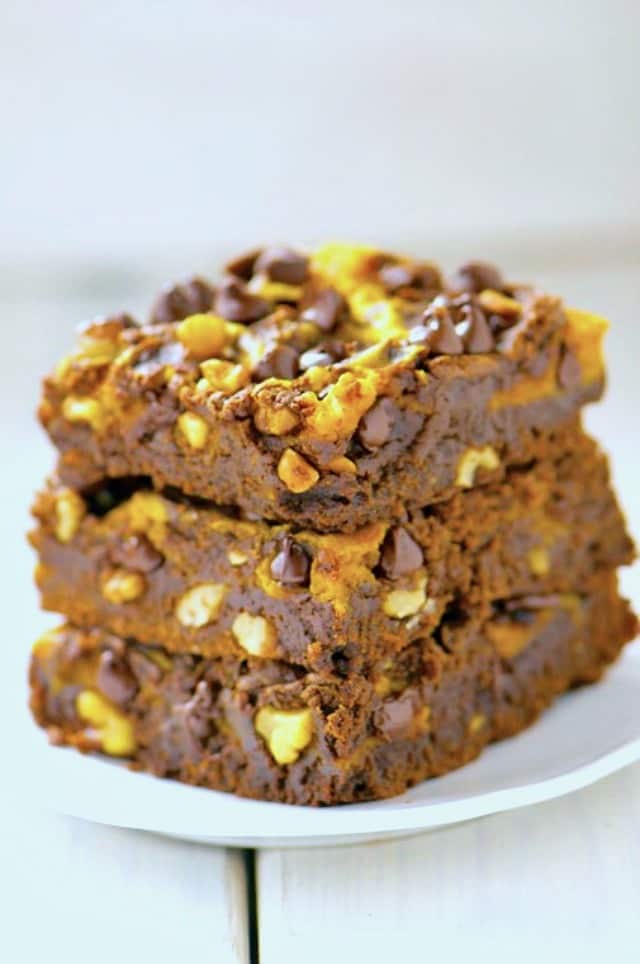 Three brownies sit one on top of another. There's lots of chocolate pieces and chopped nuts throughout.