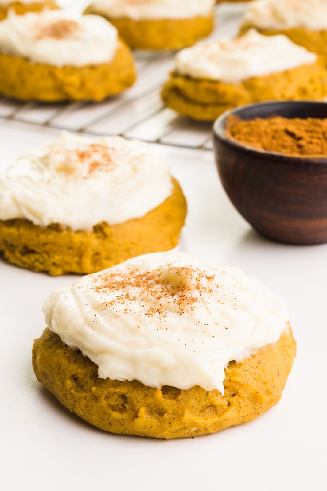 Frosted vegan pumpkin cookies sit on a white counter next to a bowl of spices. In the background is a wire rack with more cookies.