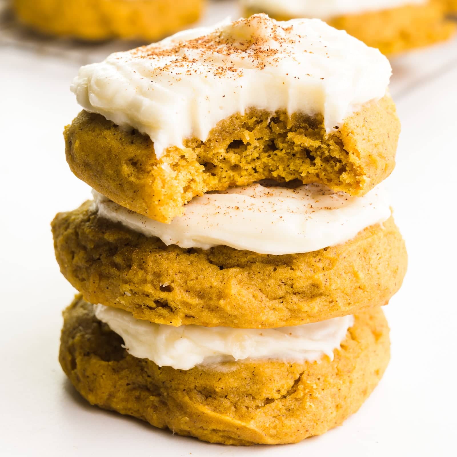 A stack of three vegan pumpkin cookies shows the top one with a bite taken out. There are more cookies in the background.