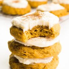 A stack of three vegan pumpkin cookies and the top one has a bite taken out. There are more cookies in the background.