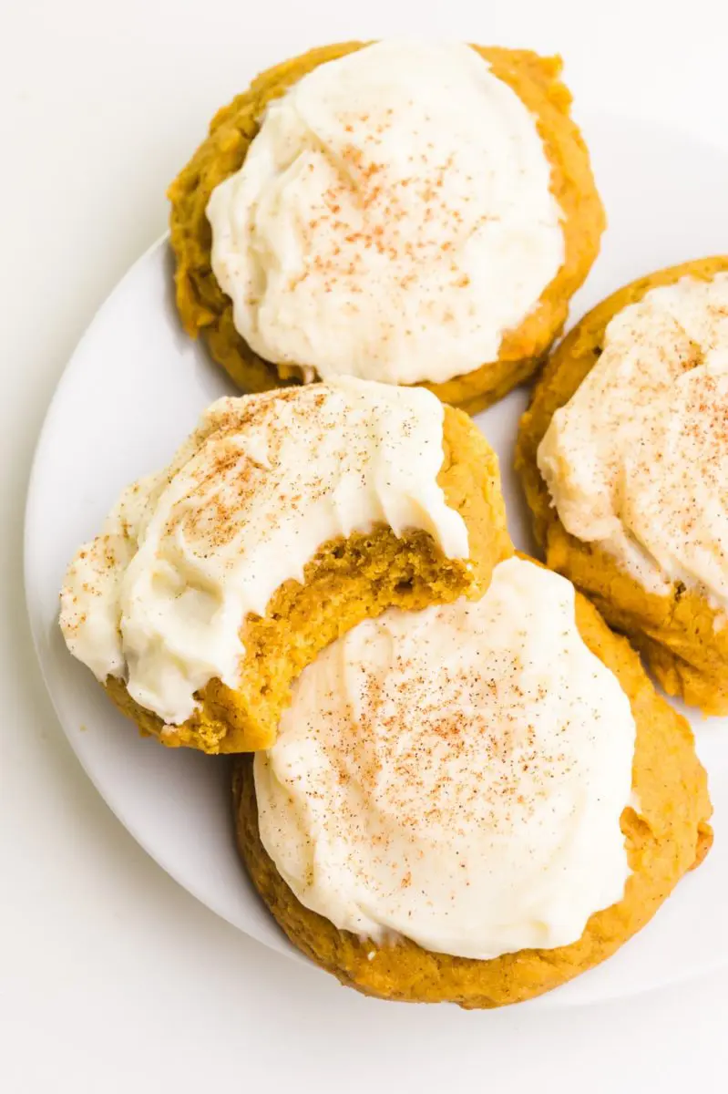 Several frosted pumpkin cookies are on a plate. One has a bite taken out.
