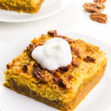 A pumpkin crisp bar sits on a white plate. It has a dollop of whipped cream on top. There's another bar on a plate and some nuts in the background.