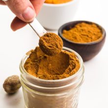 A hand holds a measuring spoon full of pumpkin pie spice over a bowlful of the spice. There's a bowl of pumpkin puree and another bowl of spice in the background.